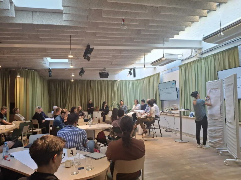 Workshop participants seated around tables listening to a panel discussion while notes are taken on a flipchart.